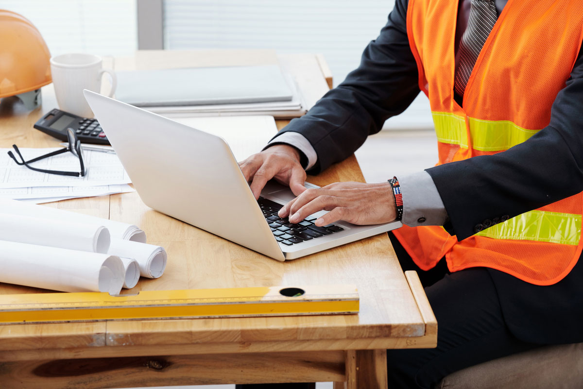 unrecognizable-man-neon-safety-vest-business-suit-sitting-desk-using-laptop-construction-document-management