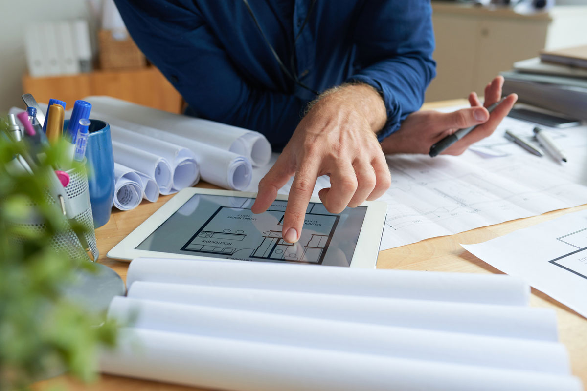 unrecognizable-man-sitting-desk-with-technical-drawings-looking-floor-plan-tablet-on-construction-takeoff